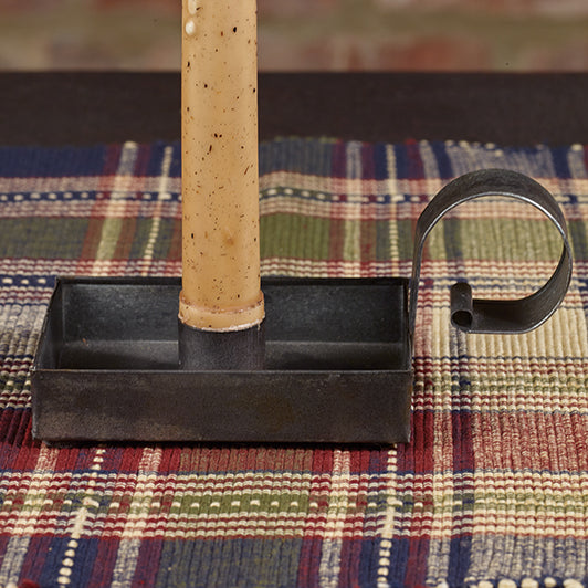 A beige colored candle placed in a black rectangular holder with a curved handle on a plaid patterned surface.