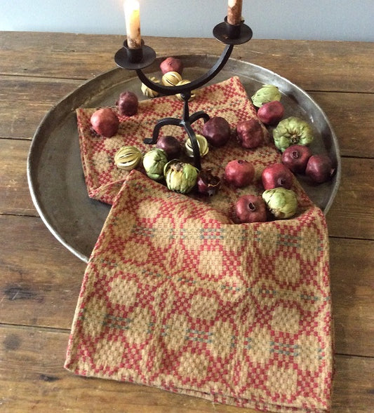 A square placemat with a red and green geometric pattern, displayed on a wooden table with a decorative candle on top, surrounded by various fruits and a black napkin ring.