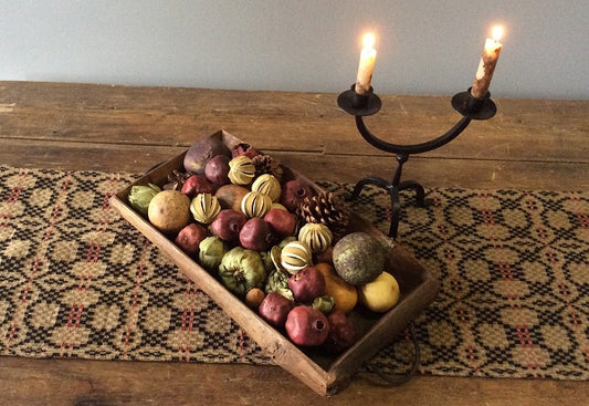A short, brown table runner with a geometric pattern, featuring a knot design, displayed on a wooden table with a decorative piece containing various fruits and vegetables.