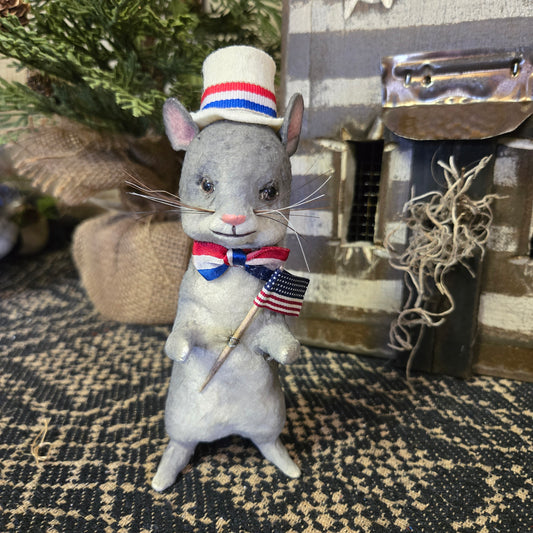 A hand-sculpted paper mache mouse with a patriotic grey, red, and blue hat and an American flag.