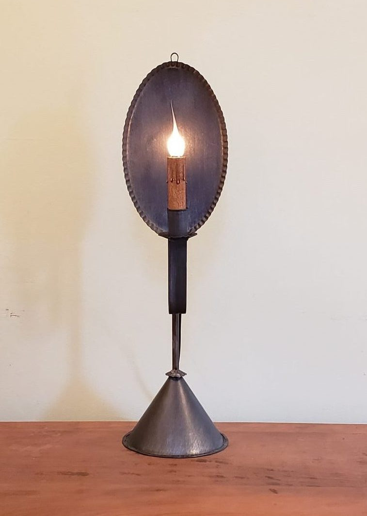 A cylindrical tin table light with one light and a black finish, placed on a wooden surface against a white wall.