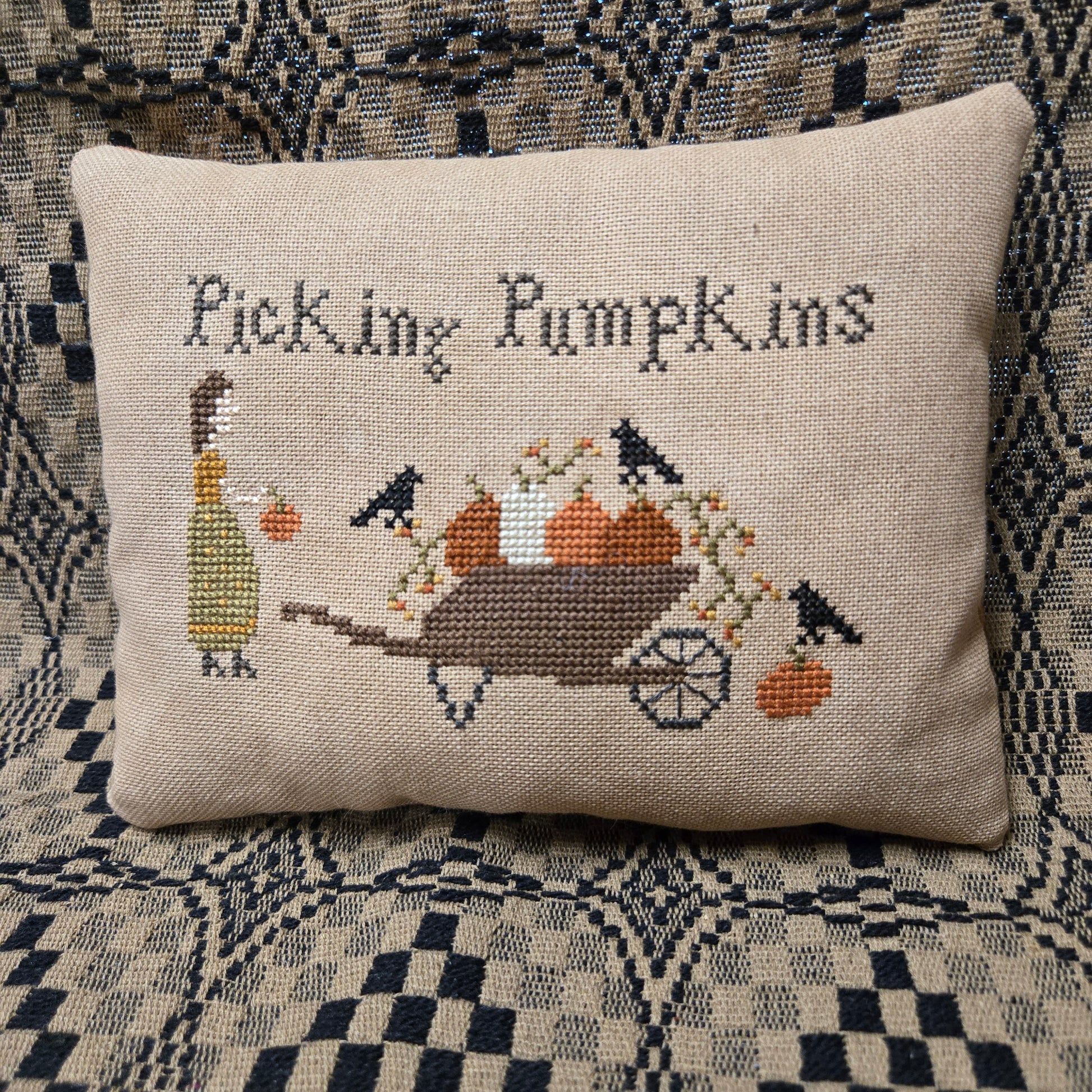 A square beige cross-stitch pillow featuring the text 'Picking Pumpkins' and a stitched illustration of a woman, pumpkins, a wheelbarrow, and birds, displayed on a patterned woven blanket.