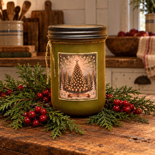 A mason jar candle filled with olive green wax, featuring a rustic metal lid and a front label depicting a glowing Christmas tree in a snowy forest. The jar sits on a wooden surface with a blurred festive background of Christmas trees and warm lights.