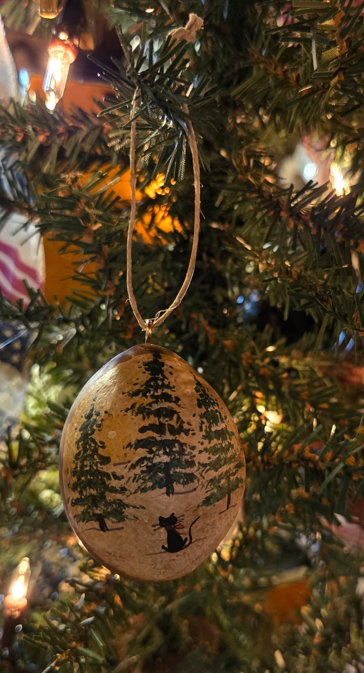A handcrafted beige and green Christmas gourd ornament with a pine tree grove design, hanging from a tree.