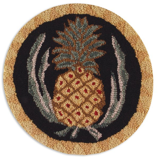 A round hooked wool chair pad featuring a central pineapple motif with gold, beige, and green colors on a black background, surrounded by a beige border.
