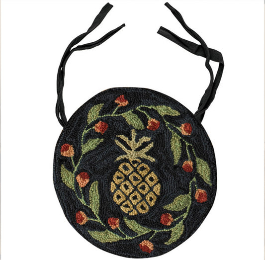 A round chair cushion with a pineapple design, featuring green pineapple, red berries, and green leaves on a black background with two black straps at the top.