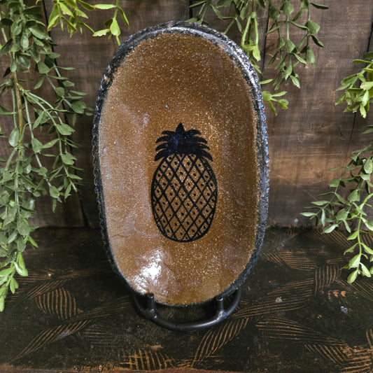 Oval-shaped brown dish with a black pineapple design on a wooden surface with green leaves.