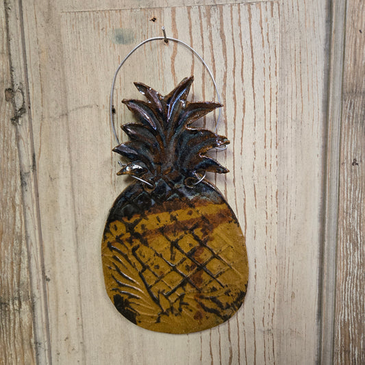 A handcrafted ceramic pineapple-shaped wall hanging with a brown and yellow design, hanging against a wooden background.