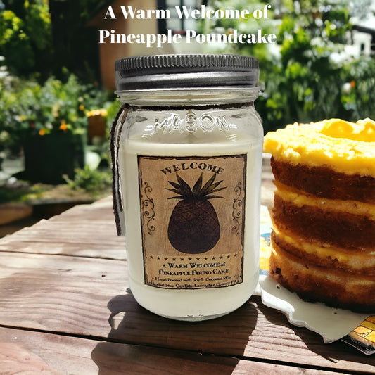 A mason jar candle with a label that reads 'Welcome' and depicts a pineapple, placed on a wooden surface with a cake in the background.