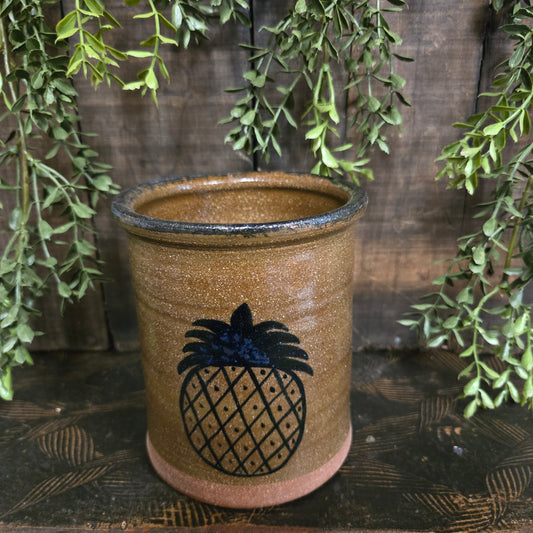 Stoneware crock with pineapple design on a wooden surface with green leaves.