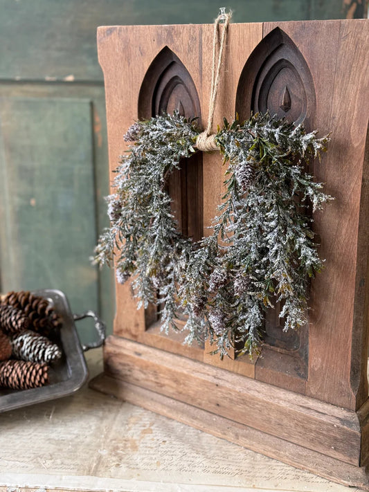 Decorative wreath hanging on a wooden panel with pinecones beside it.