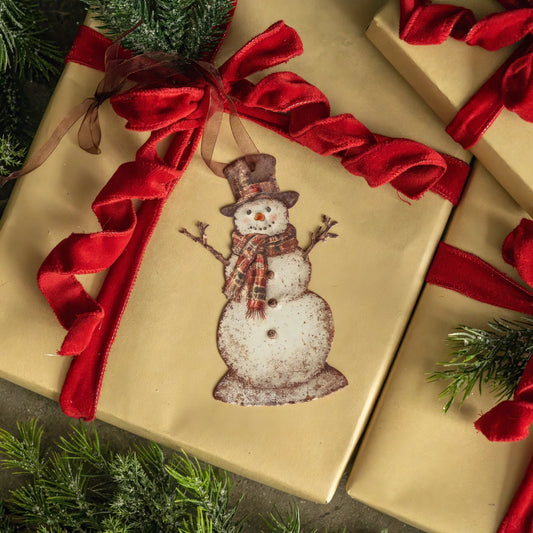 A square gift box wrapped in beige kraft paper with a large red ribbon bow and a hanging snowman ornament, surrounded by green pine branches.