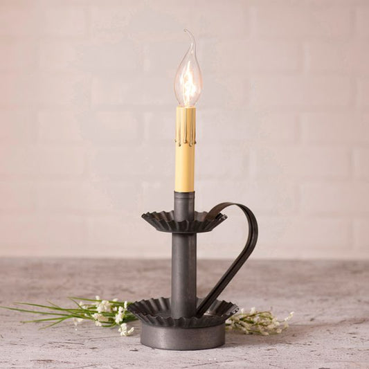 A black candlestick lamp base with a kettle-shaped design, featuring a lit candle, set against a white brick wall background with decorative dry plants.
