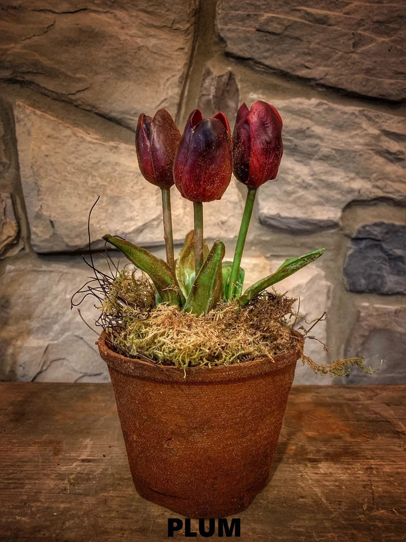 A pot with three artificial primitive style tulips, hand dipped in wax, with a brown earthy color and a textured surface, displayed on a wooden surface against a stone wall background.