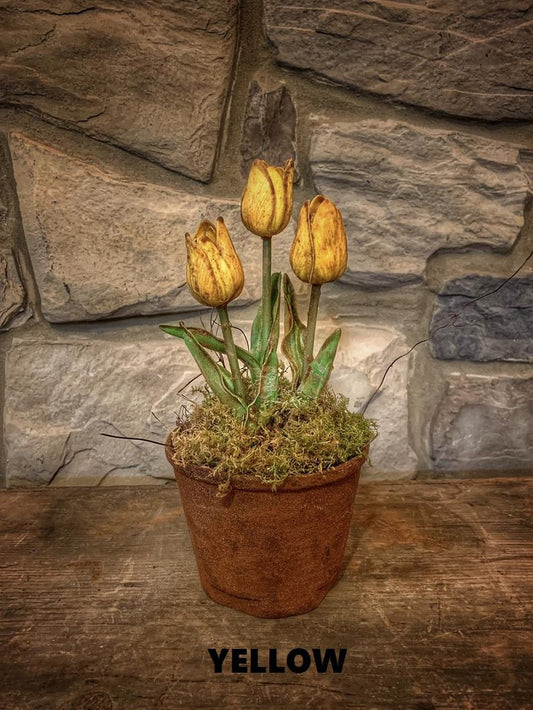 A potted plant with yellow tulips, hand dipped in wax with a rustic, primitive appearance, placed against a stone wall background.