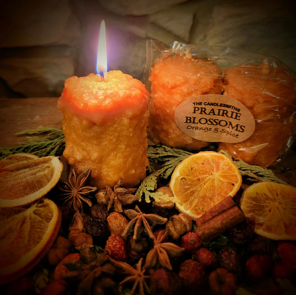 Candle with packaging labeled 'Prairie Blossoms' surrounded by oranges, cinnamon sticks, and dried herbs.