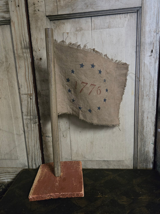 Vintage-style flag with '1776' and blue stars on a wooden stand against a rustic wooden wall.
