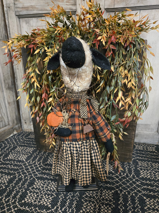 Decorative wreath with a black and white sheep figure, plaid fabric, and autumn leaves on a textured surface.