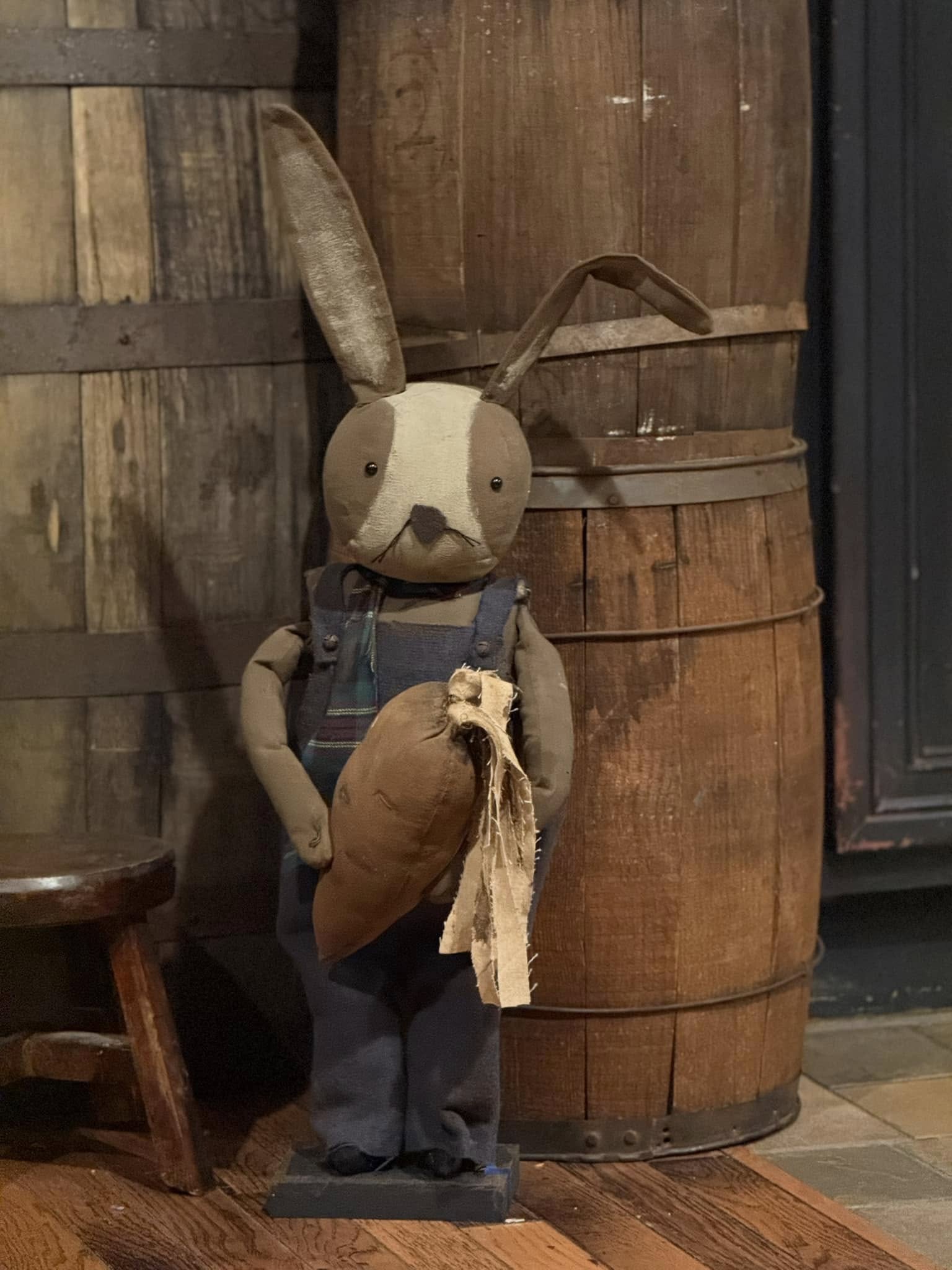 A plush toy rabbit with a grey bib, blue overalls, and a brown carrot, standing in front of wooden barrels.
