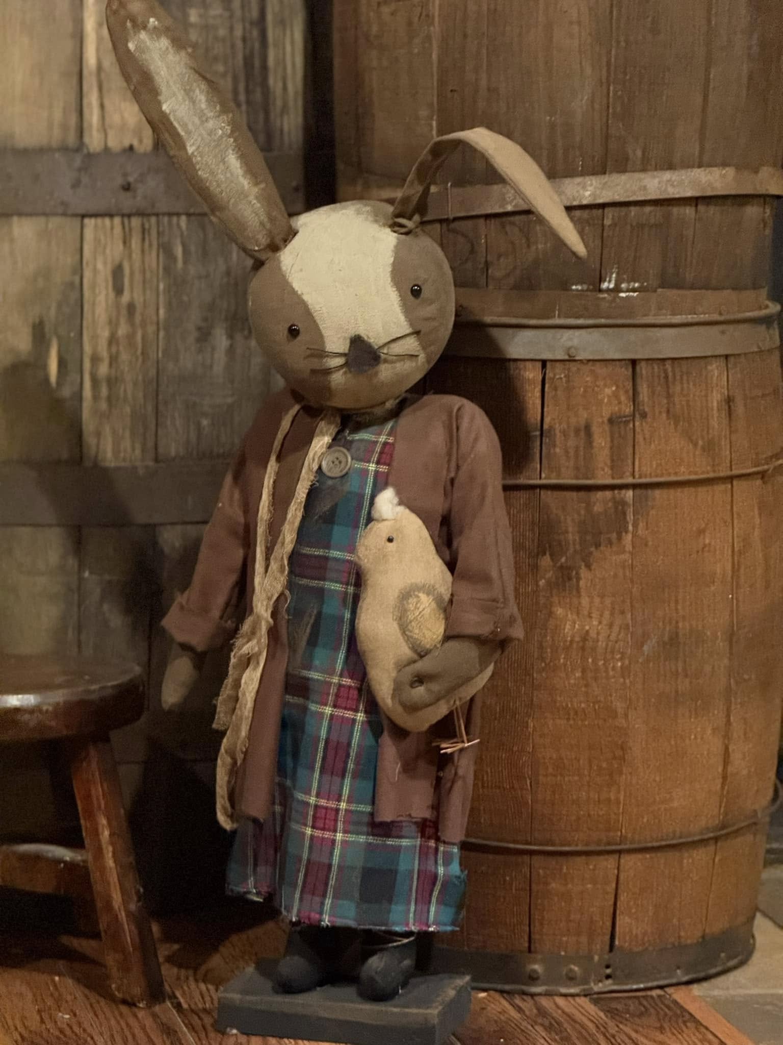 A plush toy depicting a bunny dressed in a plaid dress and a brown flannel jacket, holding a smaller plush chick, positioned in front of wooden backdrop.