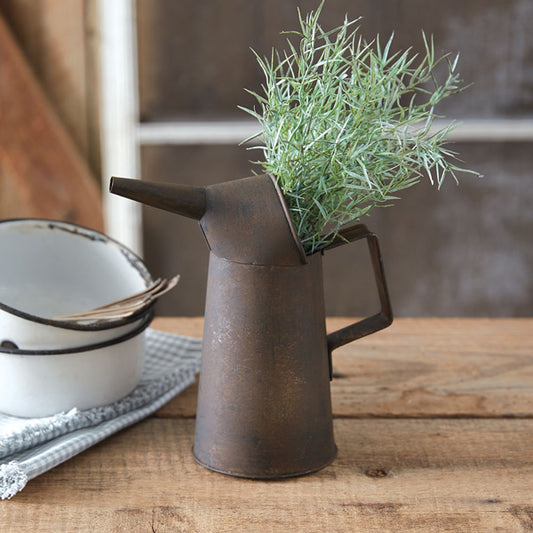 A rustic metal oil can pitcher with a spout and handle, displaying artificial greenery, placed on a wooden surface next to a white bowl.