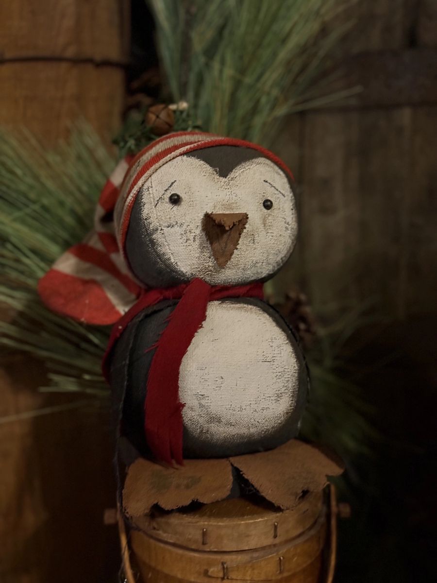 A rustic painted penguin decoration wearing a red and white striped knit hat and a red scarf, perched on a small wooden bucket with evergreen branches in the background.