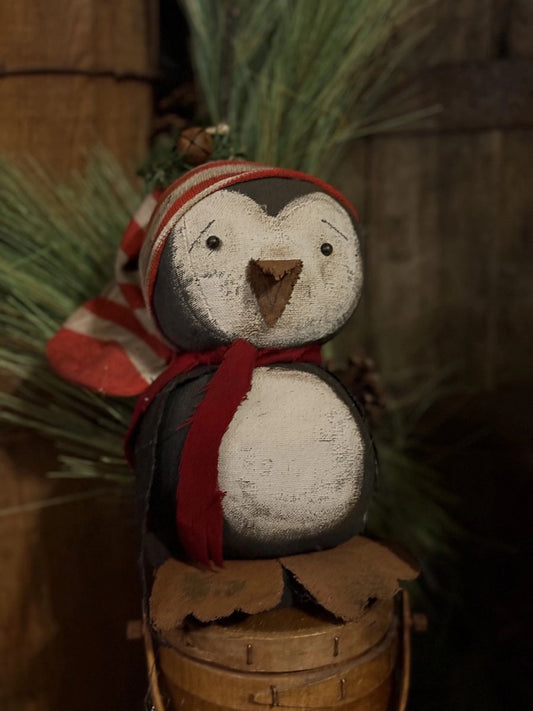 A rustic painted penguin decoration wearing a red and white striped knit hat and a red scarf, perched on a small wooden bucket with evergreen branches in the background.