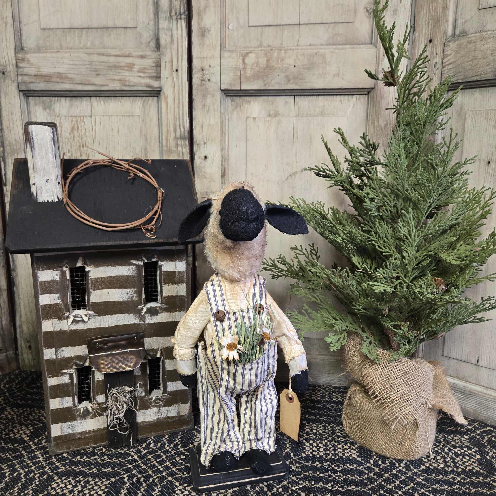 A handcrafted Primitive Baxter Sheep with a pocket of delicate flowers, dressed in a striped outfit, standing beside a decorative house and a artificial green plant.