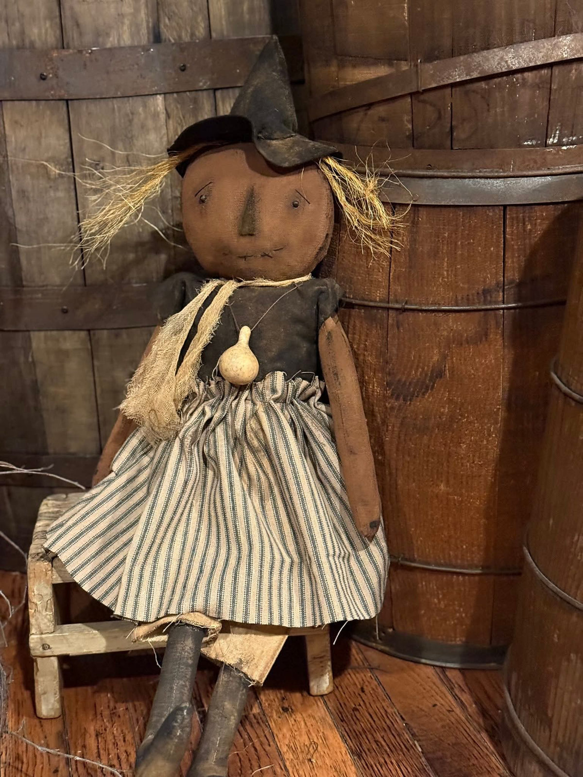Vintage-style doll with a black dress and striped apron sitting on a wooden stool against a wooden wall.