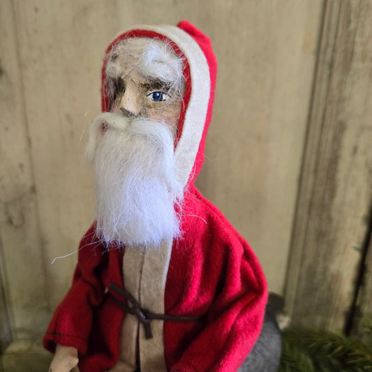 A handcrafted Primitive Belsnickel doll with a red outfit and white beard, sitting on a stool against a wooden backdrop.