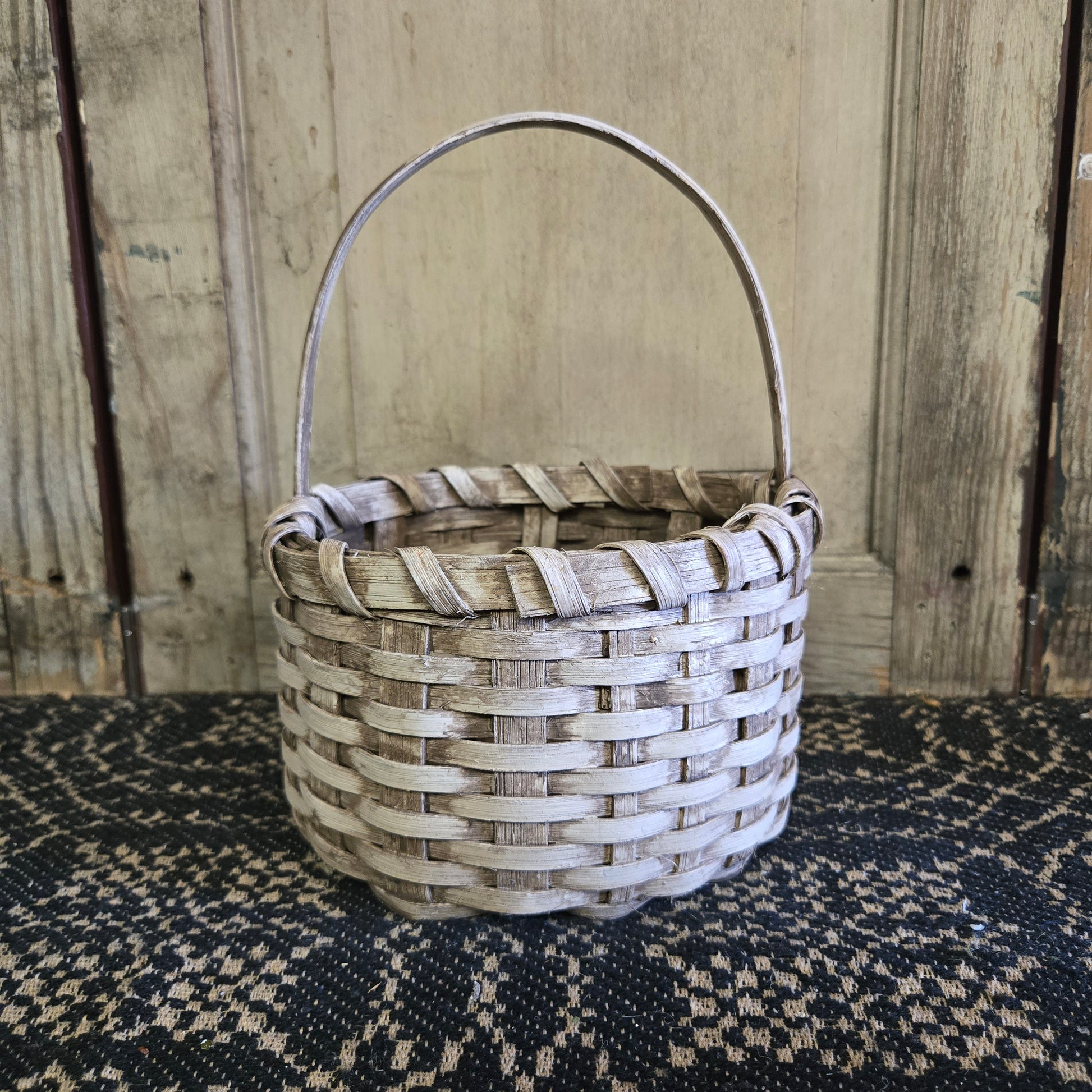 A beige, handcrafted wicker berry basket with a handle, placed on a black textured surface against a wooden backdrop.