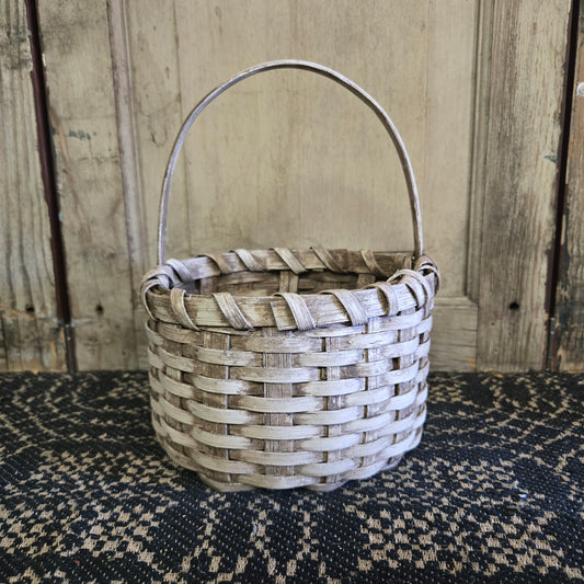 A beige, handcrafted wicker berry basket with a handle, placed on a black textured surface against a wooden backdrop.