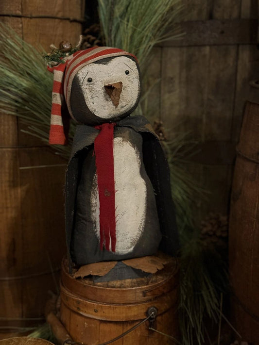 A rustic penguin decoration with a red and white striped hat, a red scarf, and a dark gray cape, displayed on a wooden barrel with pine branches in the background.