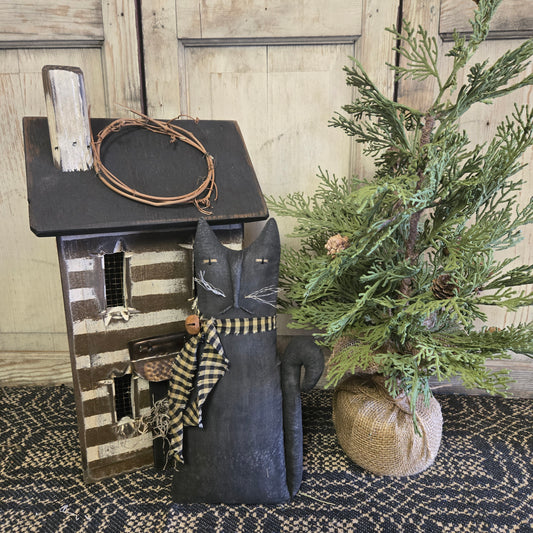 A handmade black cat figurine with a rustic bell, positioned beside a wooden structure and a green plant.