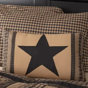 A rustic decorative throw pillow with a large black star on a beige background, styled on a bed with brown and black plaid bedding.