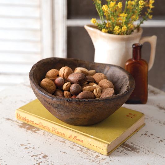 A slightly oblong, brown resin bowl filled with various nuts, placed on a yellow book, with a vase of flowers and a bottle in the background.