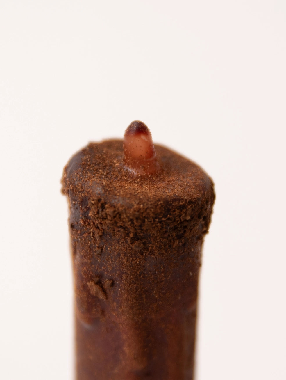 A close-up of a dark brown wax melt or candle with a textured, crumbly surface and a small amber-colored wick, set against a plain white background.