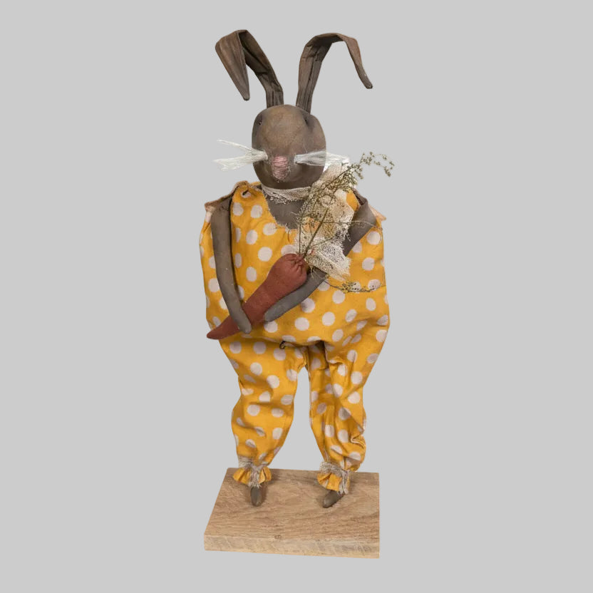 Vintage doll in a yellow polka dot dress standing on a wooden base against a rustic wooden wall.