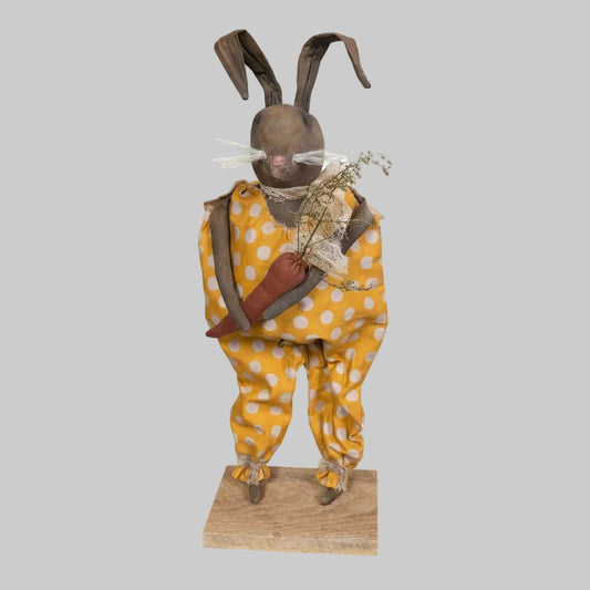 Vintage doll in a yellow polka dot dress standing on a wooden base against a rustic wooden wall.