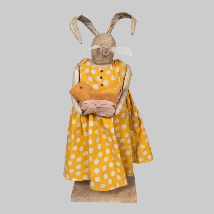 Stuffed animal in a yellow dress with white polka dots on a wooden base.