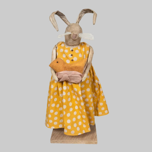 Stuffed animal in a yellow dress with white polka dots on a wooden base.