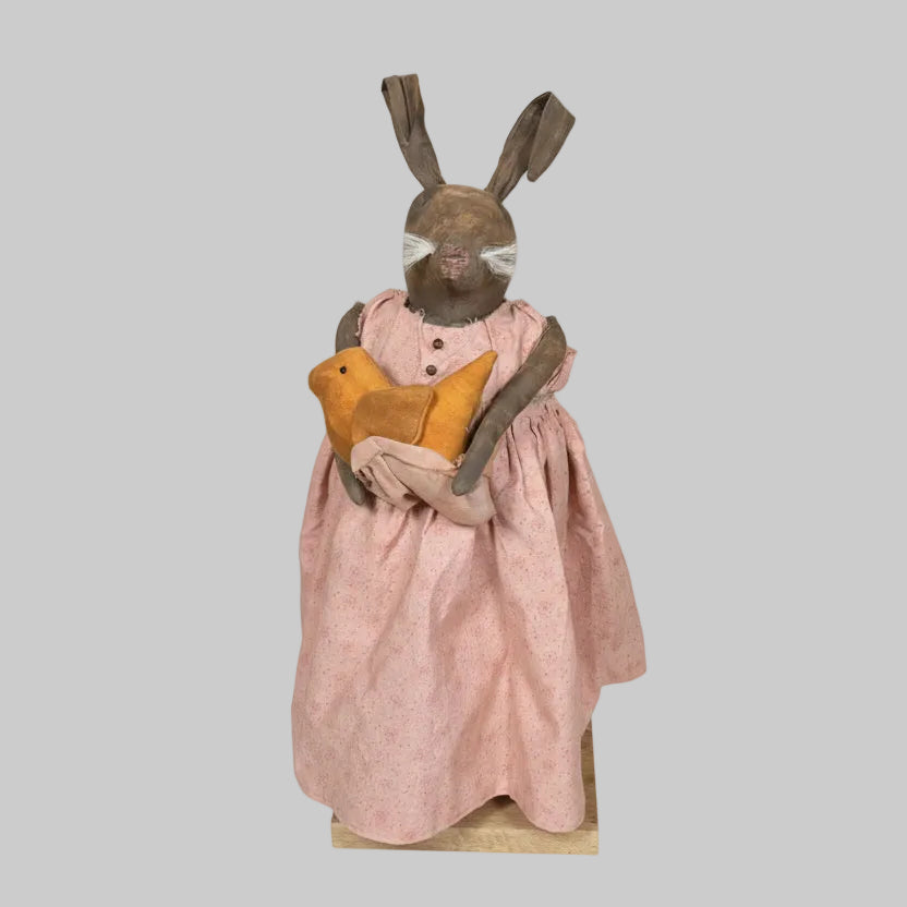 Bunny wearing a pink dress with a yellow apron standing in front of a wooden wall.