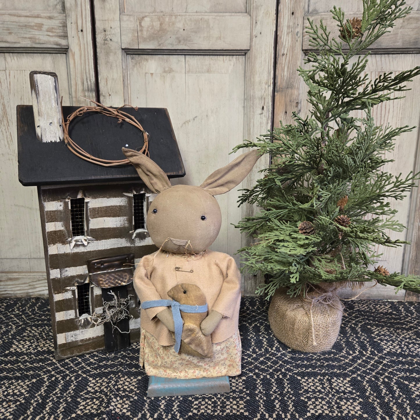A beige-colored bunny figurine holding a chick, placed on a decorative mat with a green plant and a birdhouse in the background.
