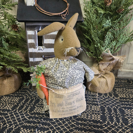 A handmade plush toy resembling a beige-colored bunny with a carrot and apron, displayed in a rustic setting with greenery and a wooden crate background.
