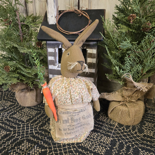 A handmade primitive bunny with a carrot and apron, displayed in a rustic setting with greenery and a wooden backdrop.