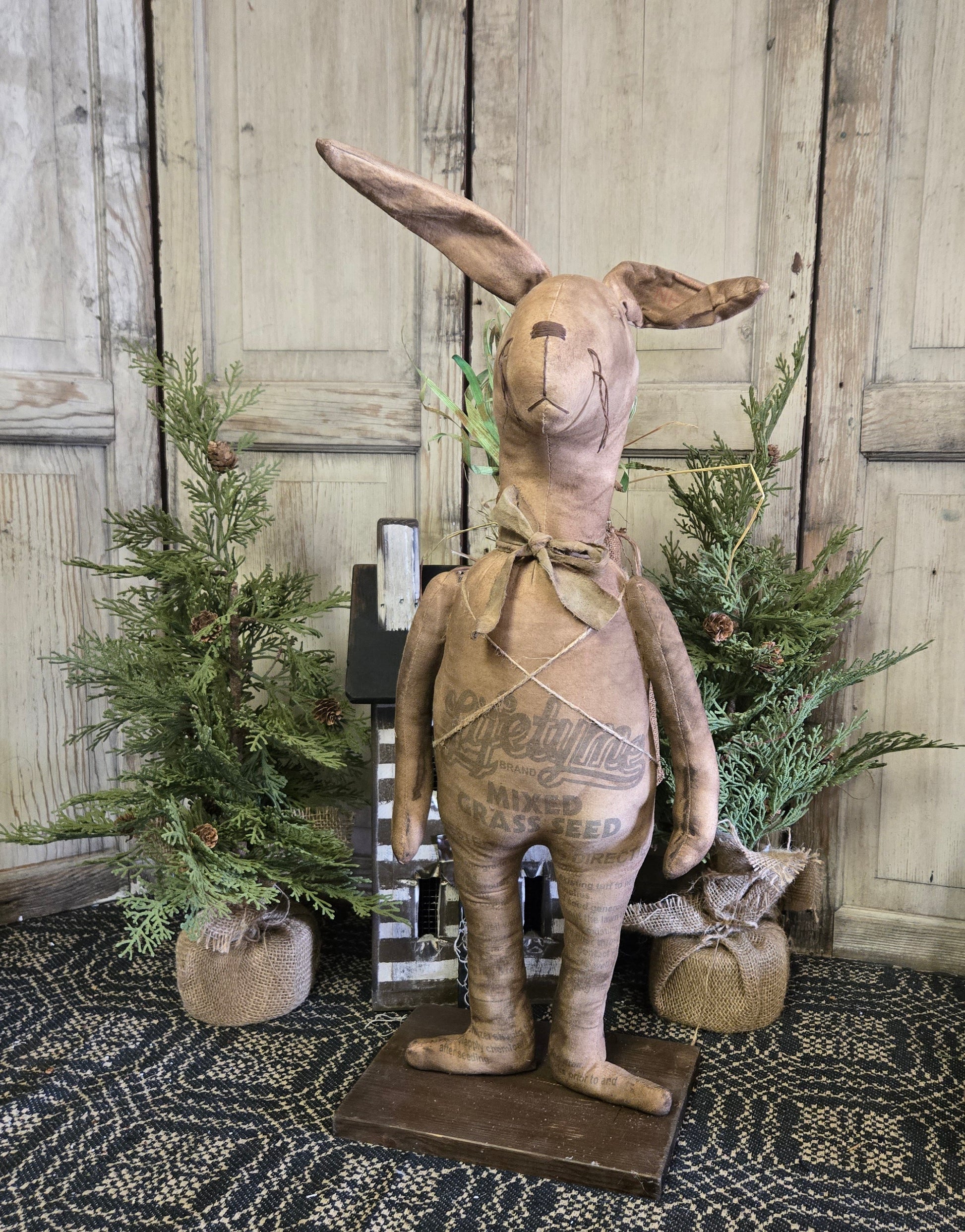 A beige fabric doll representing a primitive bunny with a carrot sack on its back, displayed against a rustic backdrop.