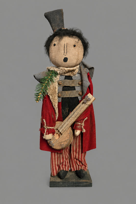 A rustic folk art figurine of a singing man playing a banjo, dressed in a red coat, striped pants, and a top hat, standing on a dark table against a grey background.