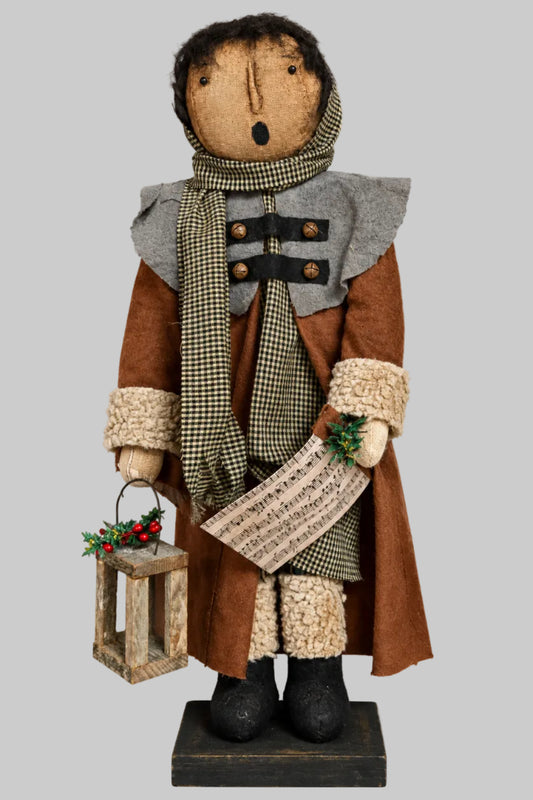 A rustic handmade rag doll figurine dressed in a brown coat, gray shawl, and checkered scarf, holding a lantern and paper, set against a solid gray background.