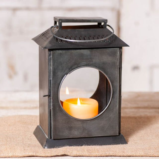 A black metal carriage lantern with two round glass windows, a lit candle inside casting a warm glow, placed on a burlap surface against a textured background.
