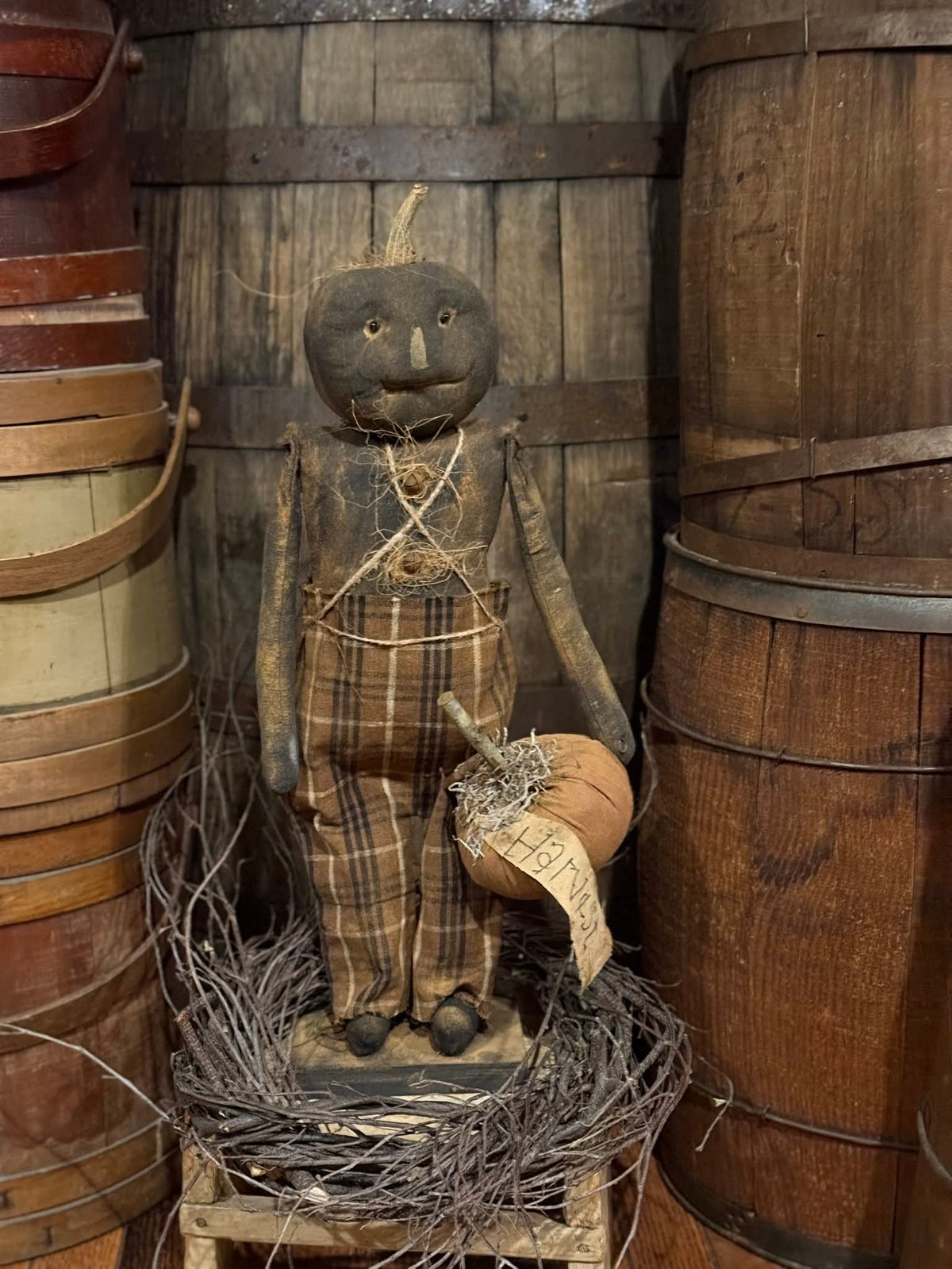 Vintage-style scarecrow figure with plaid overalls and pumpkin head, surrounded by wooden barrels.